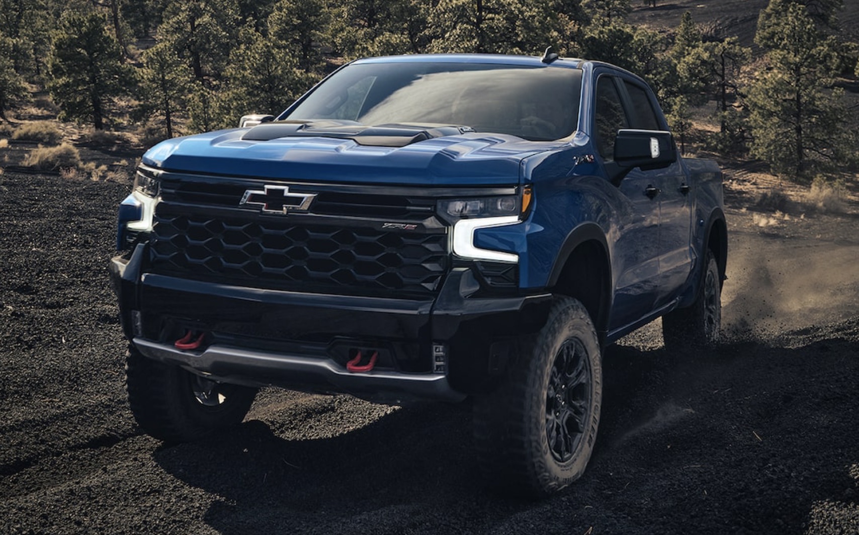 Blue Chevrolet Silverado Driving Up A Hill, Off Road With Lots Of Trees In The Background